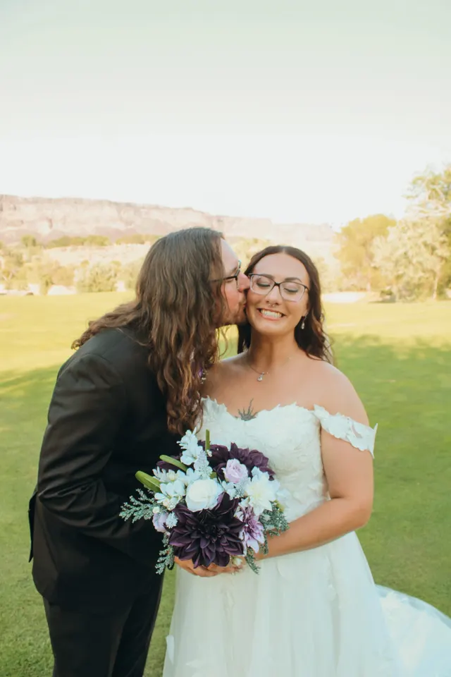 Groom kissing the bride on the cheek on a sunlit lawn