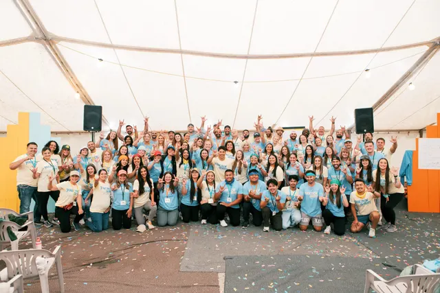 About a hundred coworkers in matching blue and cream branded t-shirts, hands in the air under a white event tent with confetti across the floor