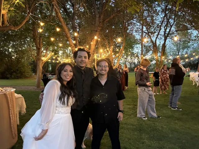 Newlyweds posing with DJ Manny under string lights at an outdoor reception