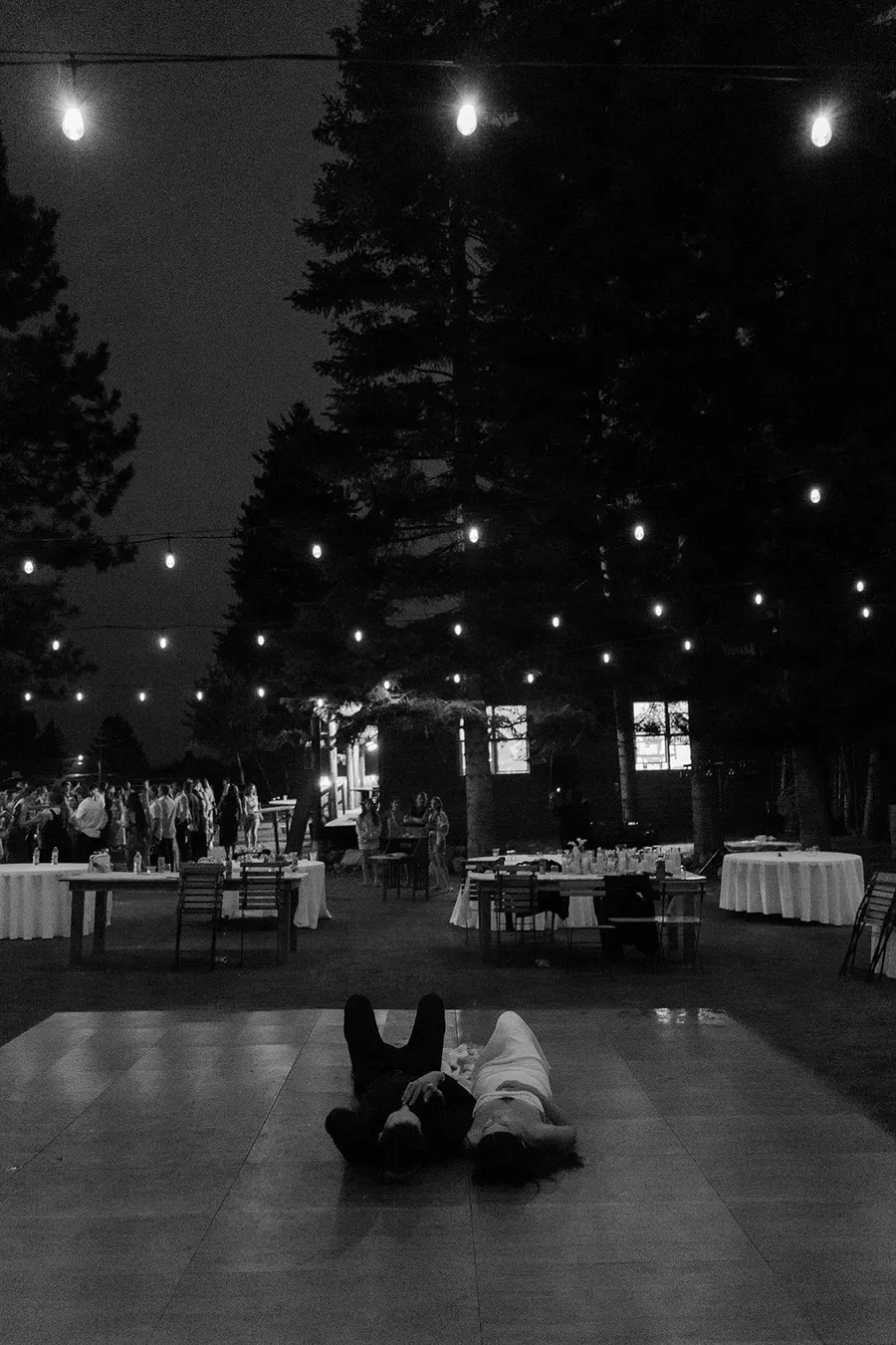 The newlyweds lying together on an empty dance floor at the end of the night, string lights overhead and the last guests packing up behind them