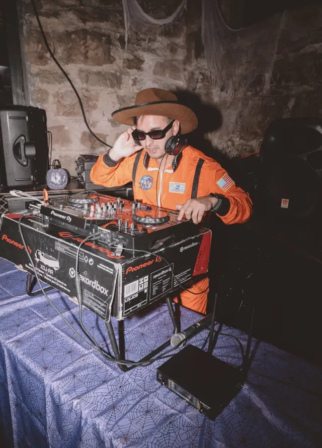 DJ Manny in a hat and orange jacket behind the decks at a stone-walled venue