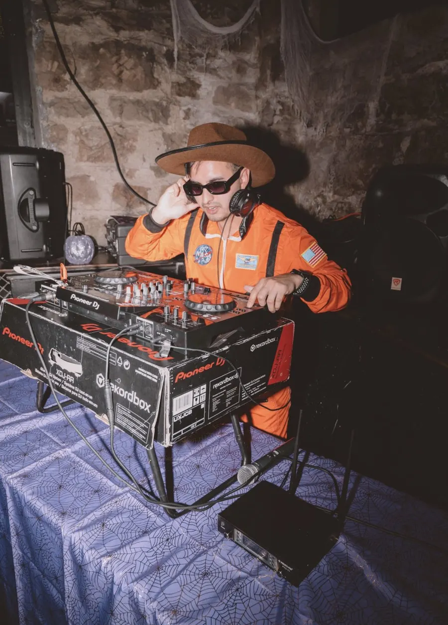 DJ Manny in a hat and orange jacket behind the decks at a stone-walled venue