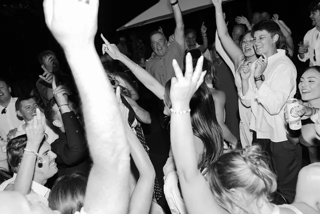 A packed dance floor at night in black and white, guests with their hands up singing along shoulder to shoulder