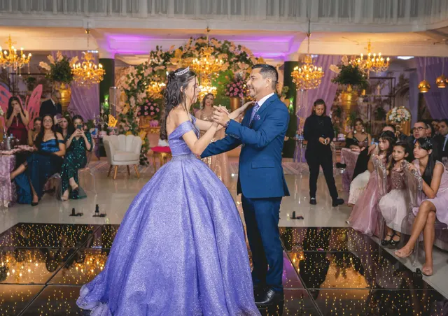 A quinceanera in a lavender ball gown dancing the vals with her father on a lit dance floor, her guests watching from tables under chandeliers