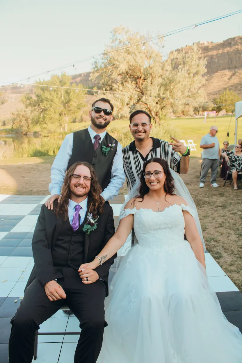 A newly married couple seated together on a checkered dance floor at their outdoor wedding, with a groomsman and DJ Manny standing behind them and canyon hills beyond