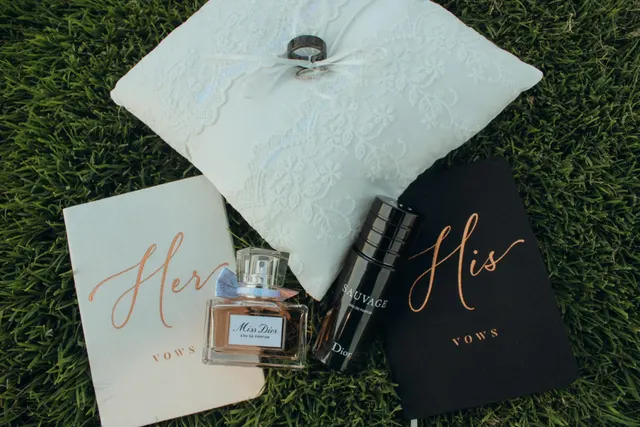 Her and His vow books laid out on grass beside two perfume bottles and a lace ring pillow holding the wedding bands