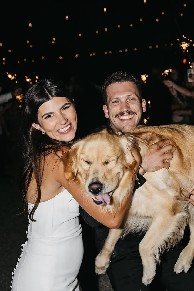 A newlywed couple laughing as they hold up their golden retriever during their sparkler send-off, warm string lights glowing behind them